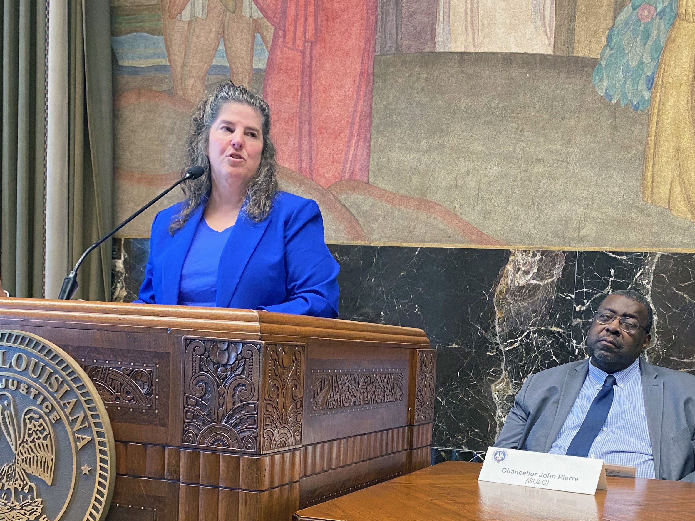Administrator Liz Ryan standing behind a podium. Seated to her left is SULC Chancellor John Pierre