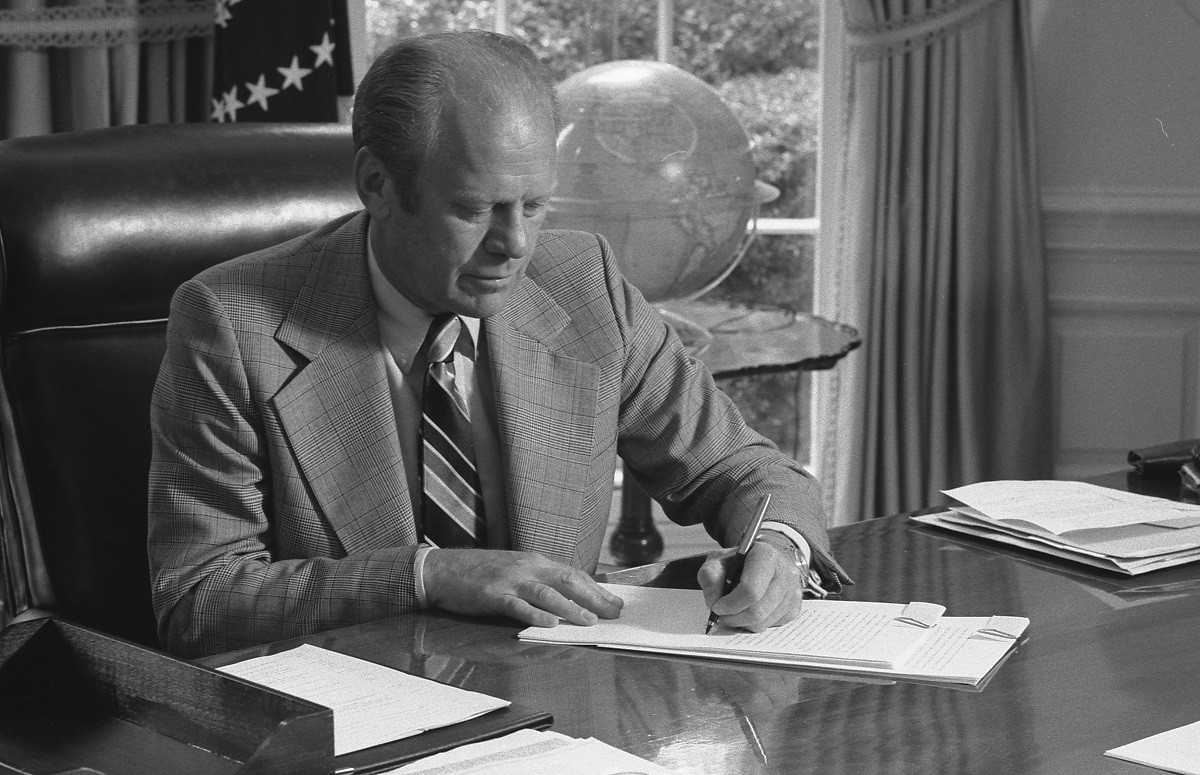 A black and white photograph shows President Gerald Ford sitting at the Resolute Desk in the Oval Office. He's wearing a plaid suit and striped tie, intently writing on papers. The American flag is visible in the background, and a globe sits on a nearby table. Ford's focused posture captures him in a moment of presidential duties, likely during his 1974-1977 term.